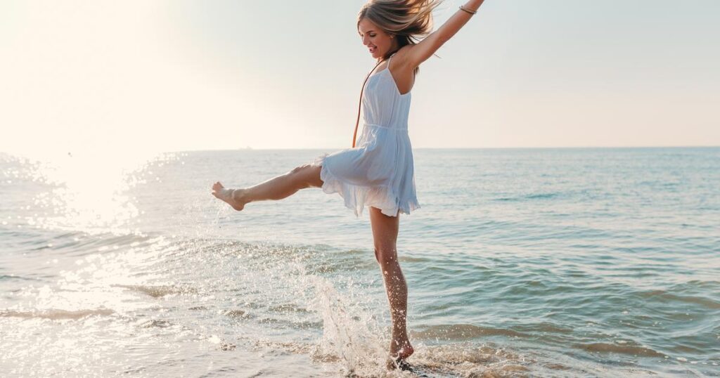 A woman in a white dress joyfully jumps into the ocean, embodying the spirit of "What would you wear at the beach?