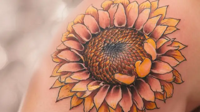 A woman's shoulder adorned with a sunflower tattoo, showcasing a unique style that highlights intricate details and vibrant colors.